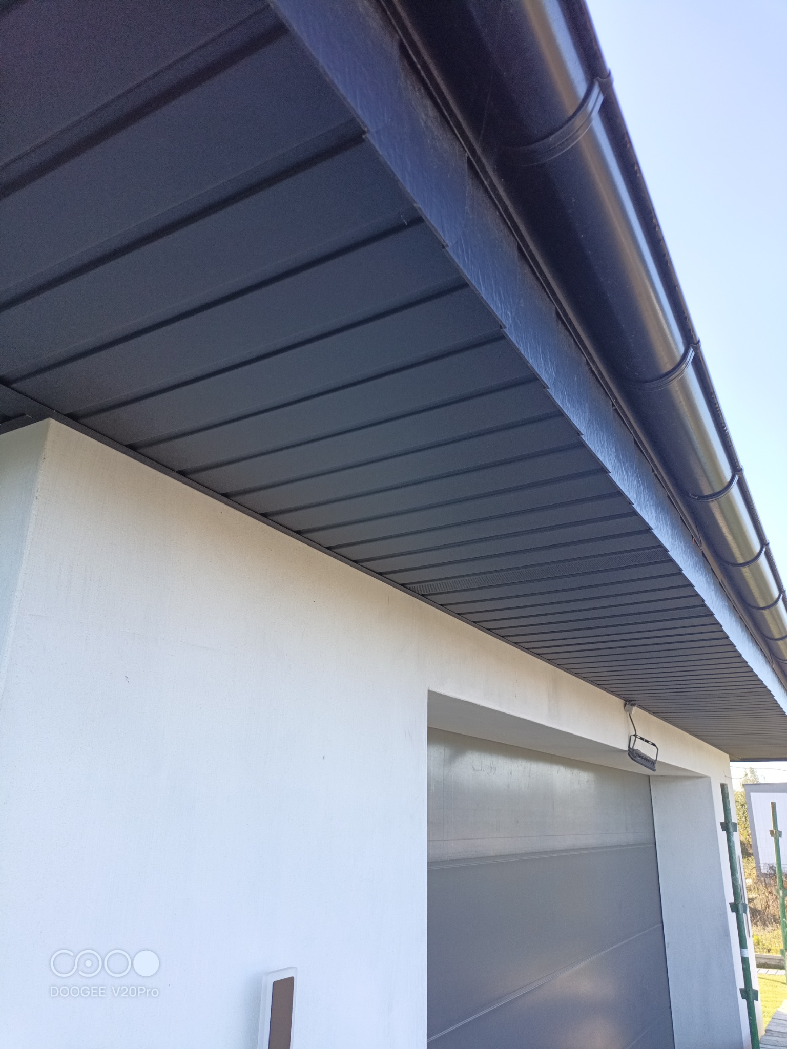 Szara podbitka dachowa z blachy trapezowej i rynna w kolorze grafitowym zamontowane przy białej elewacji budynku.