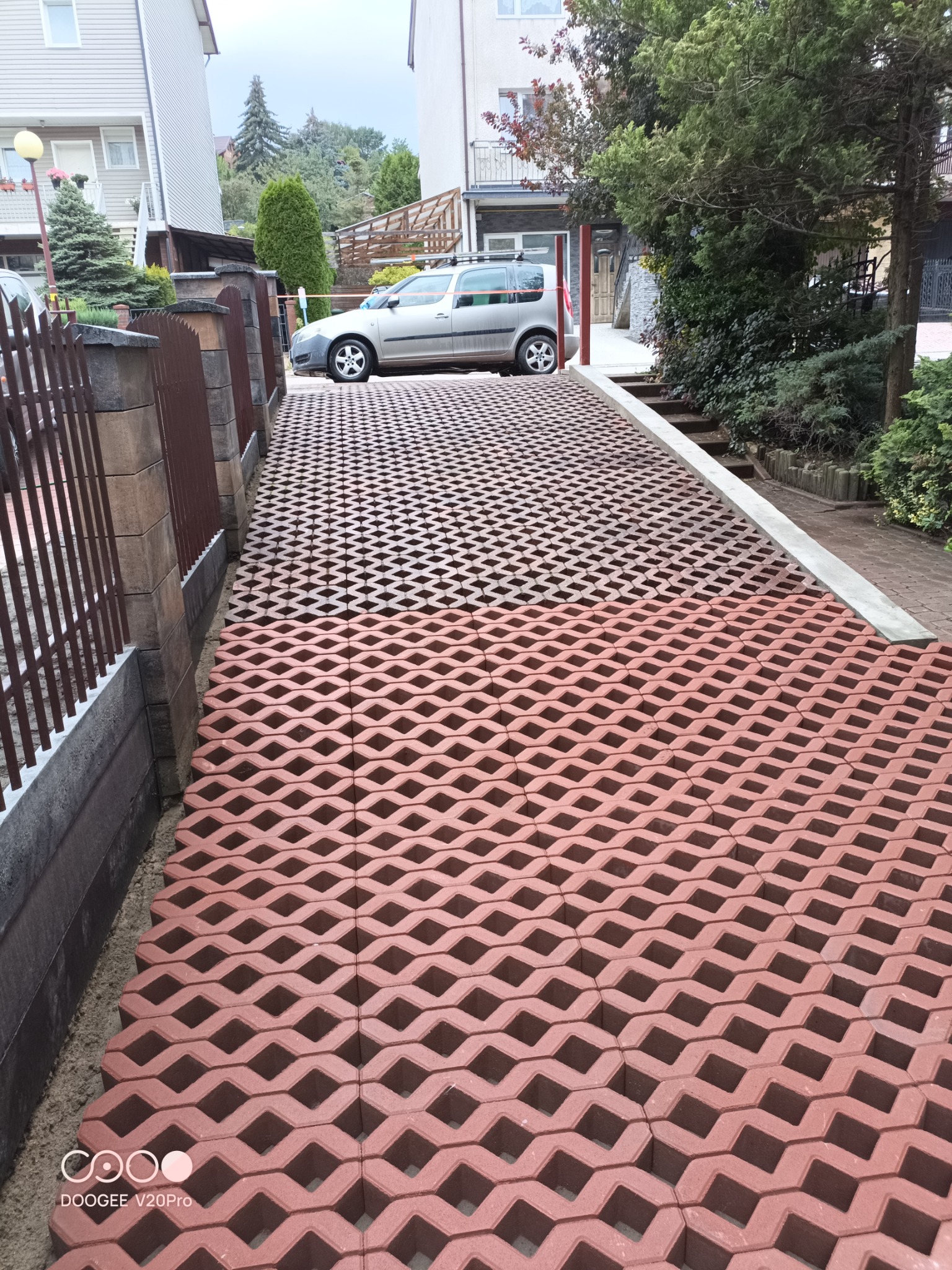 Ułożona, ażurowa nawierzchnia podjazdu z czerwonej kostki na tle częściowo widocznego ogrodzenia, domu i zaparkowanego samochodu.