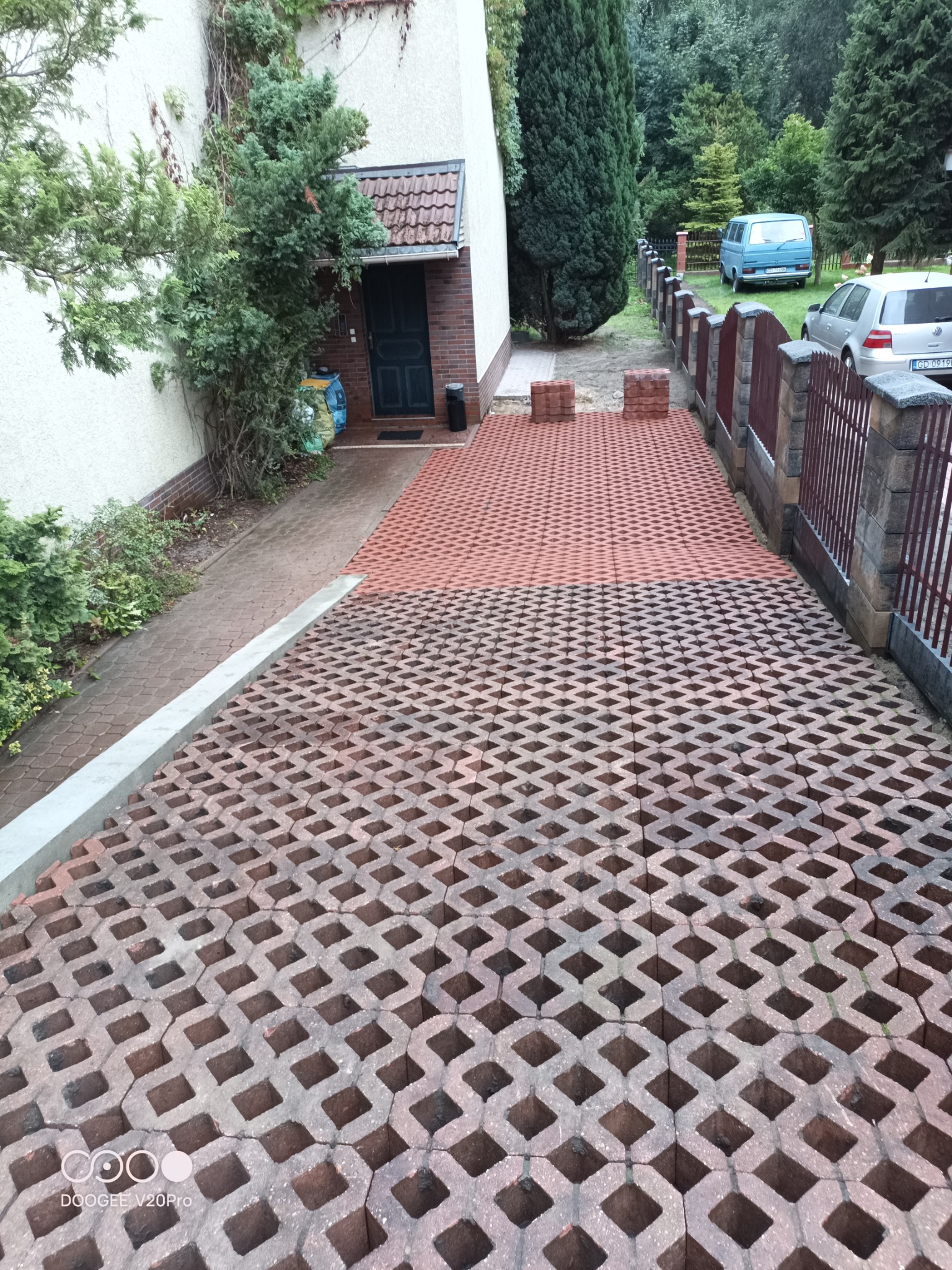 Częściowo ułożona nawierzchnia z ażurowych, czerwono-brązowych płyt betonowych na podjeździe przed domem, widoczny fragment chodnika z kostki, ogrodzenie z metalowymi przęsłami i słupkami z cegły.