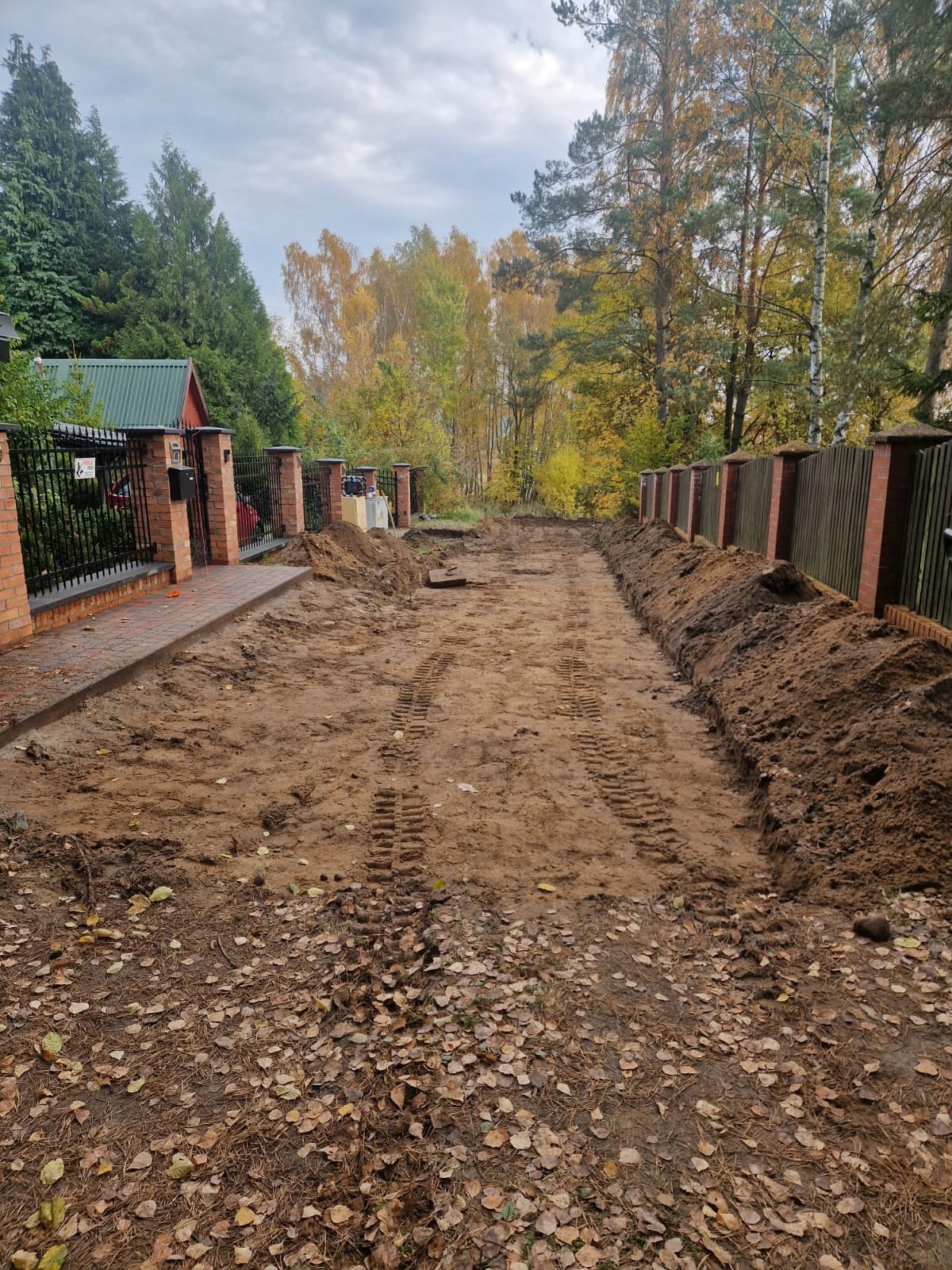 Przygotowanie terenu pod montaż ogrodzenia: wykopane rowy wzdłuż linii ogrodzenia, ślady opon na piasku, widoczny fragment istniejącego ogrodzenia z ceglanymi słupkami i metalowymi przęsłami...