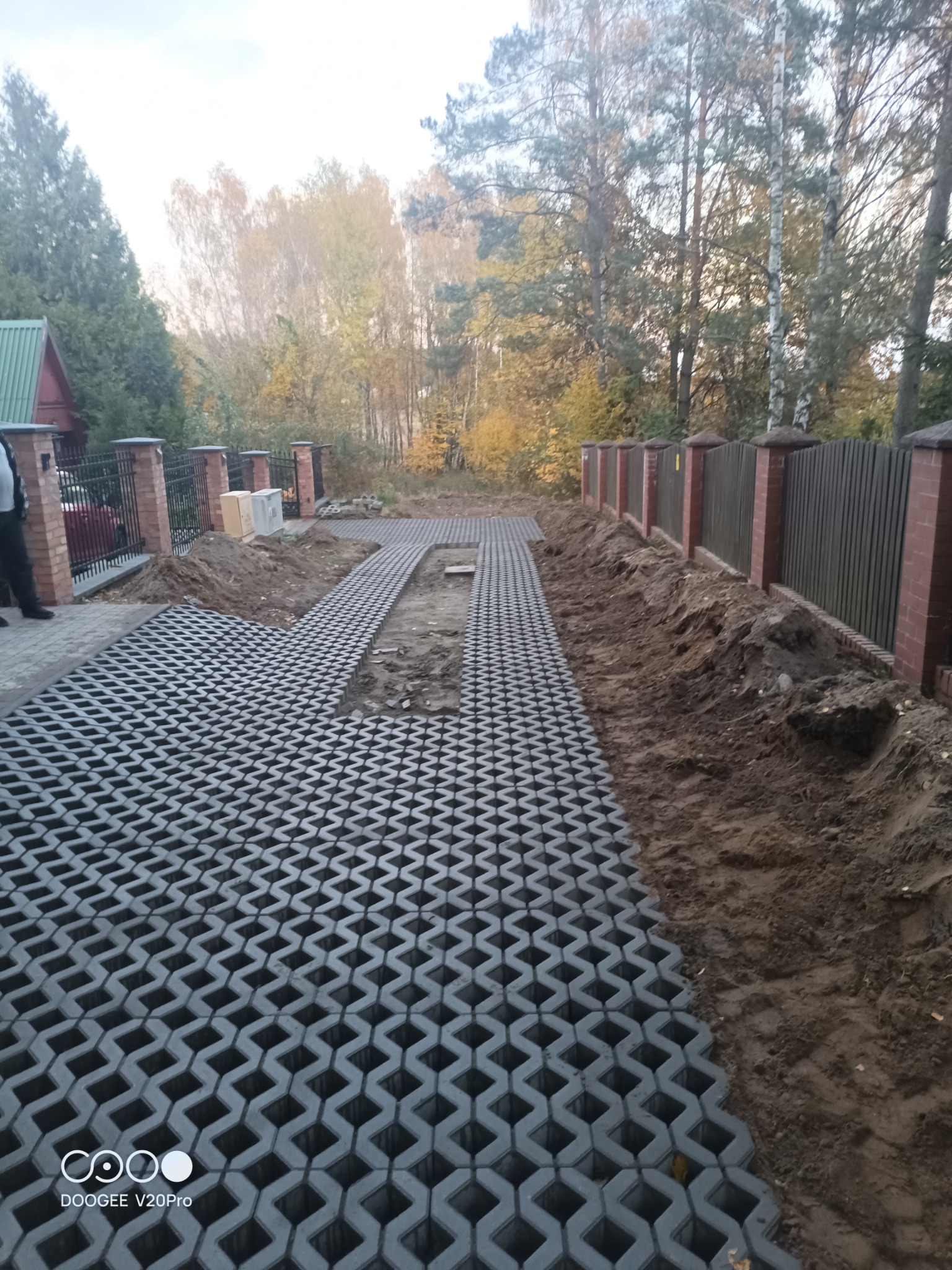Układanie szarej ażurowej nawierzchni na podjeździe, widoczny fragment ogrodzenia z ceglanymi słupkami i drewnianymi przęsłami, ziemia po bokach, w tle drzewa.