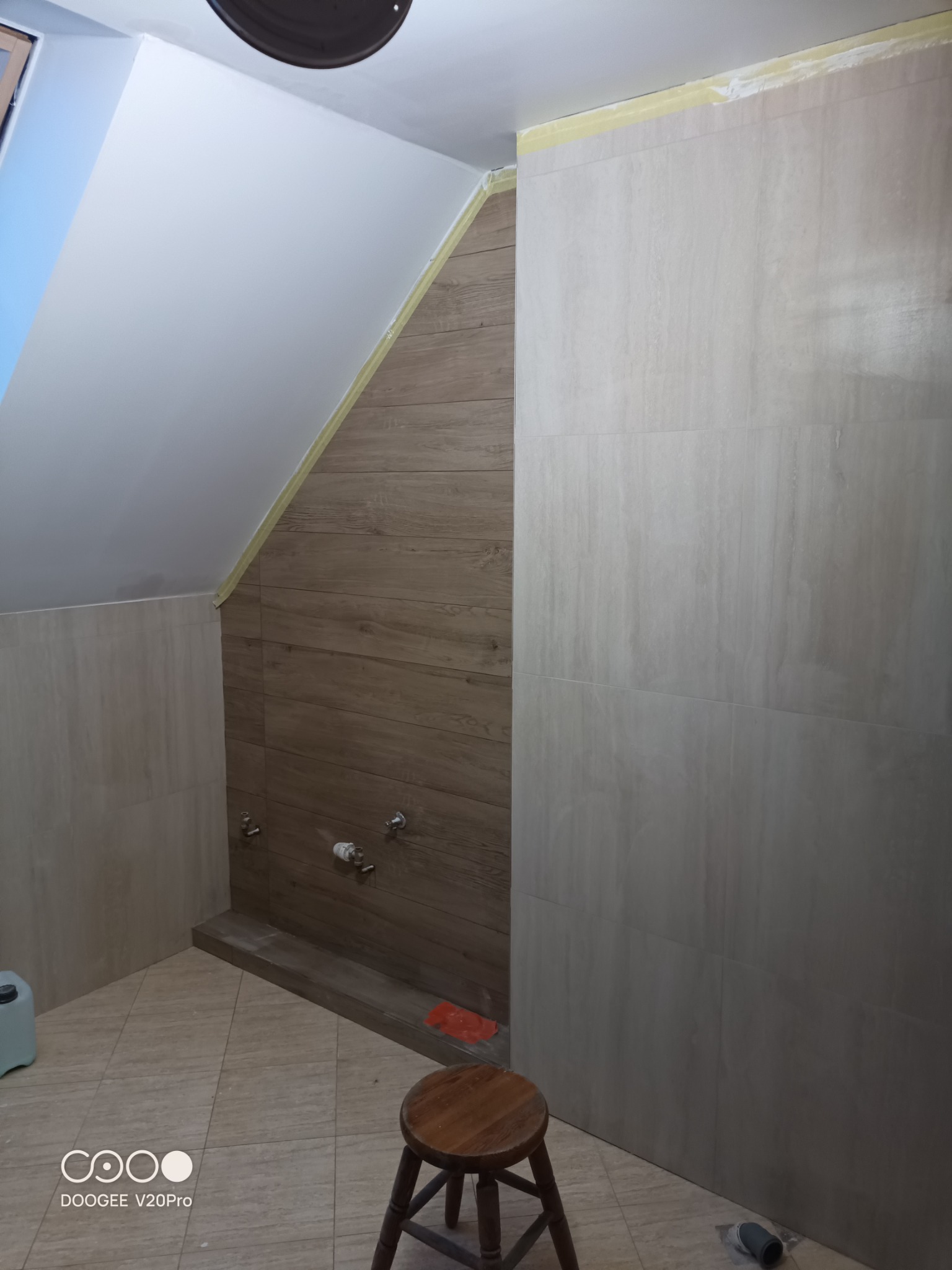 Wykończona łazienka na poddaszu z beżowymi i drewnopodobnymi płytkami, widoczne przygotowane przyłącza wodne, żółta taśma malarska zabezpieczająca krawędzie i niski stołek.