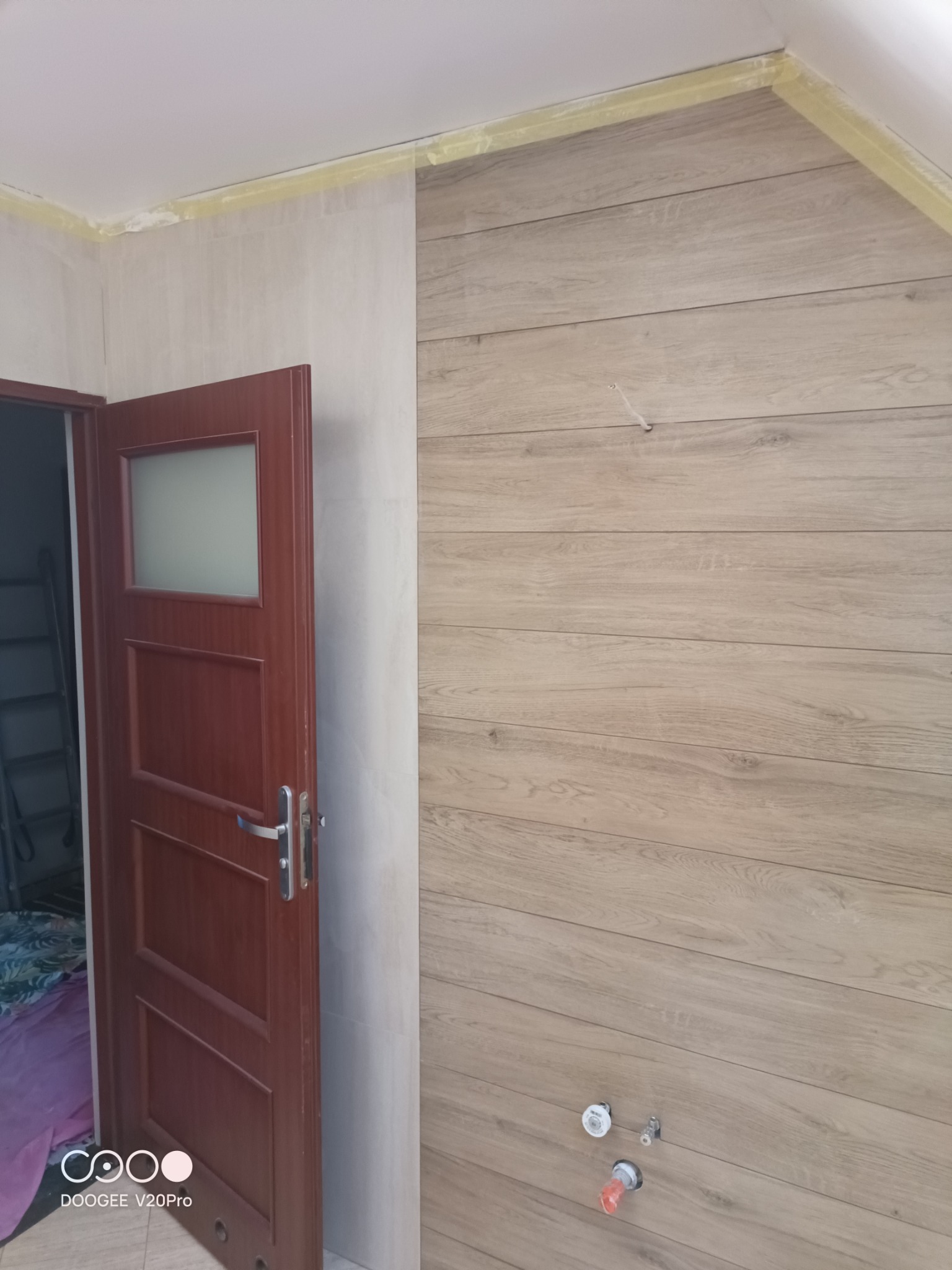 Wnętrze łazienki w trakcie remontu: drewnopodobne panele ścienne, zabezpieczone taśmą malarską przy suficie, częściowo otwarta, ciemnobrązowa, przeszklona wewnętrzna stolarka drzwiowa...