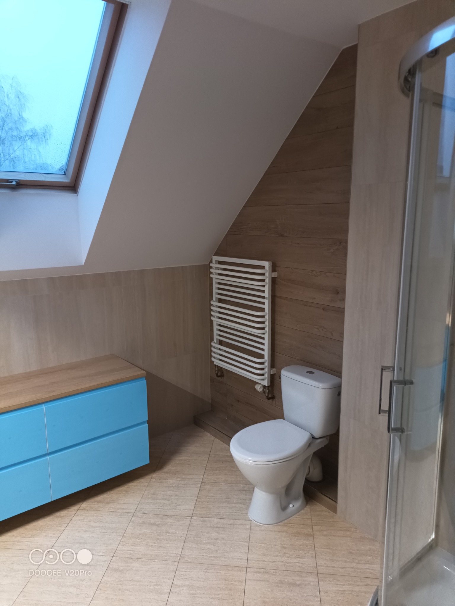 Jasna łazienka na poddaszu ze skośnym oknem dachowym, turkusową szafką pod umywalkę, grzejnikiem drabinkowym i kabiną prysznicową.