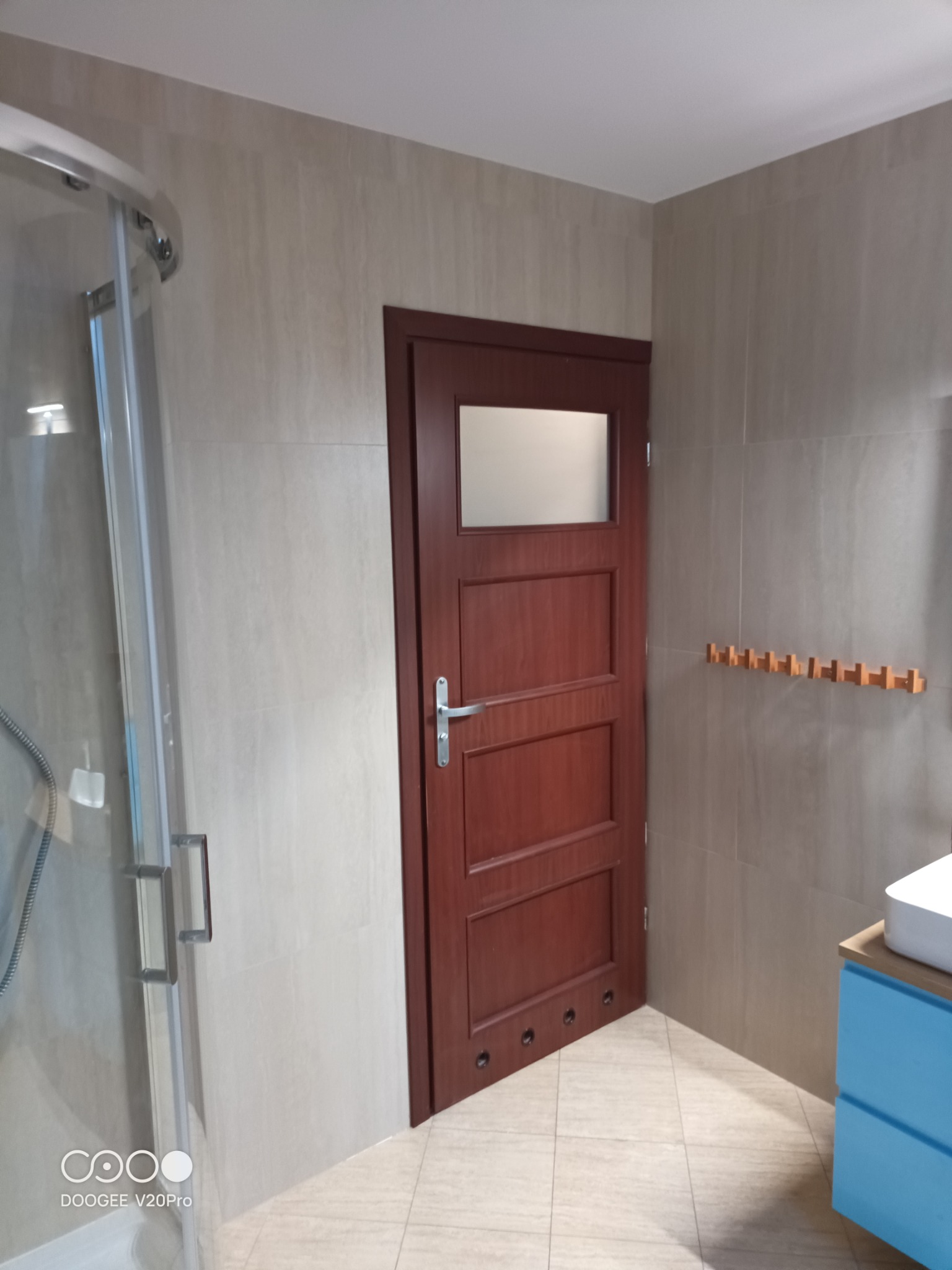 Nowoczesna łazienka z kabiną prysznicową, drewnianymi drzwiami z przeszkleniem, błękitną szafką pod umywalką i wieszakiem na ręczniki w kolorze drewna.