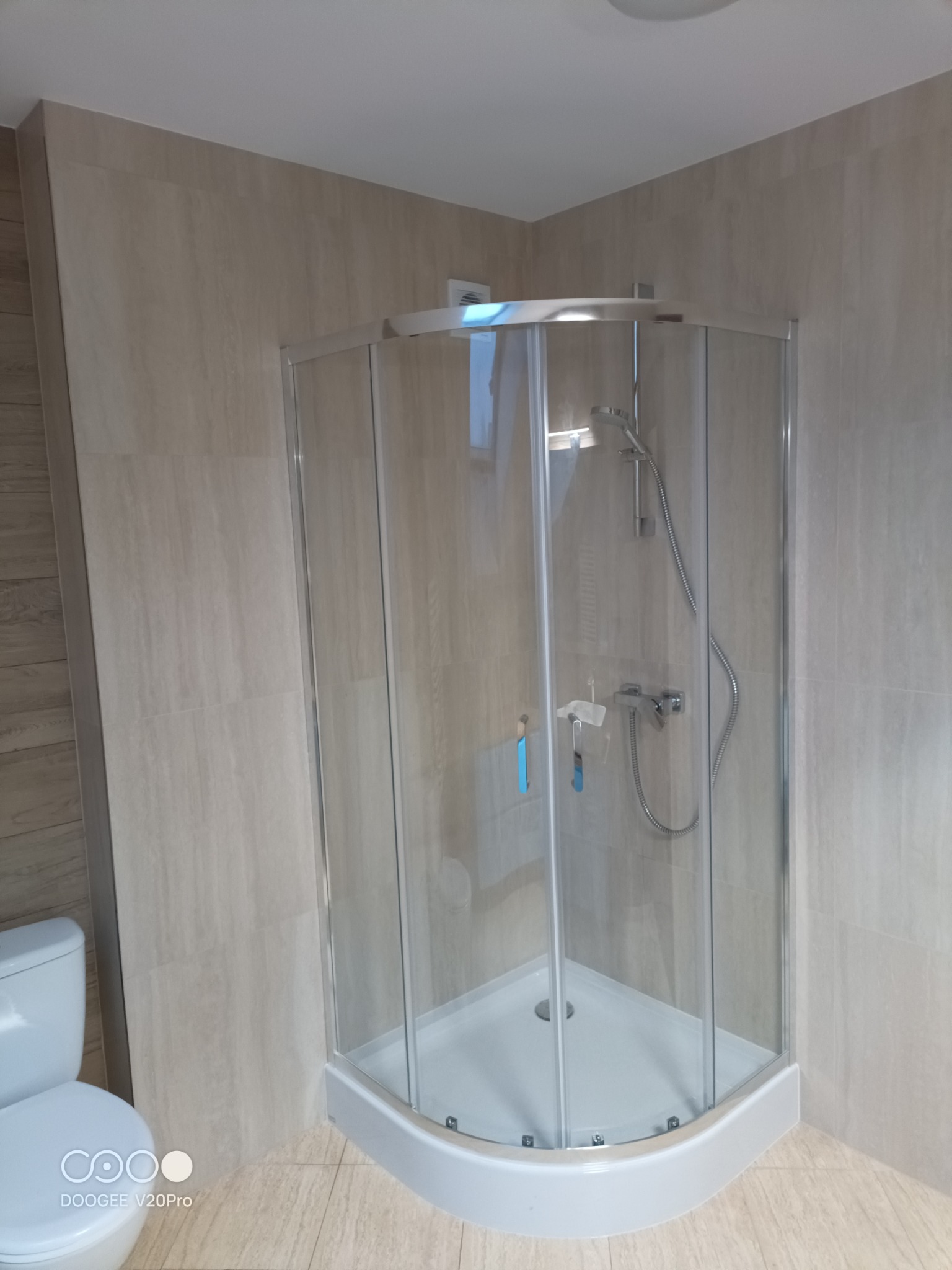 Narożna kabina prysznicowa z chromowanymi profilami i białą brodzikiem, przylegająca do toalety, wykończona płytkami imitującymi drewno w odcieniach beżu.