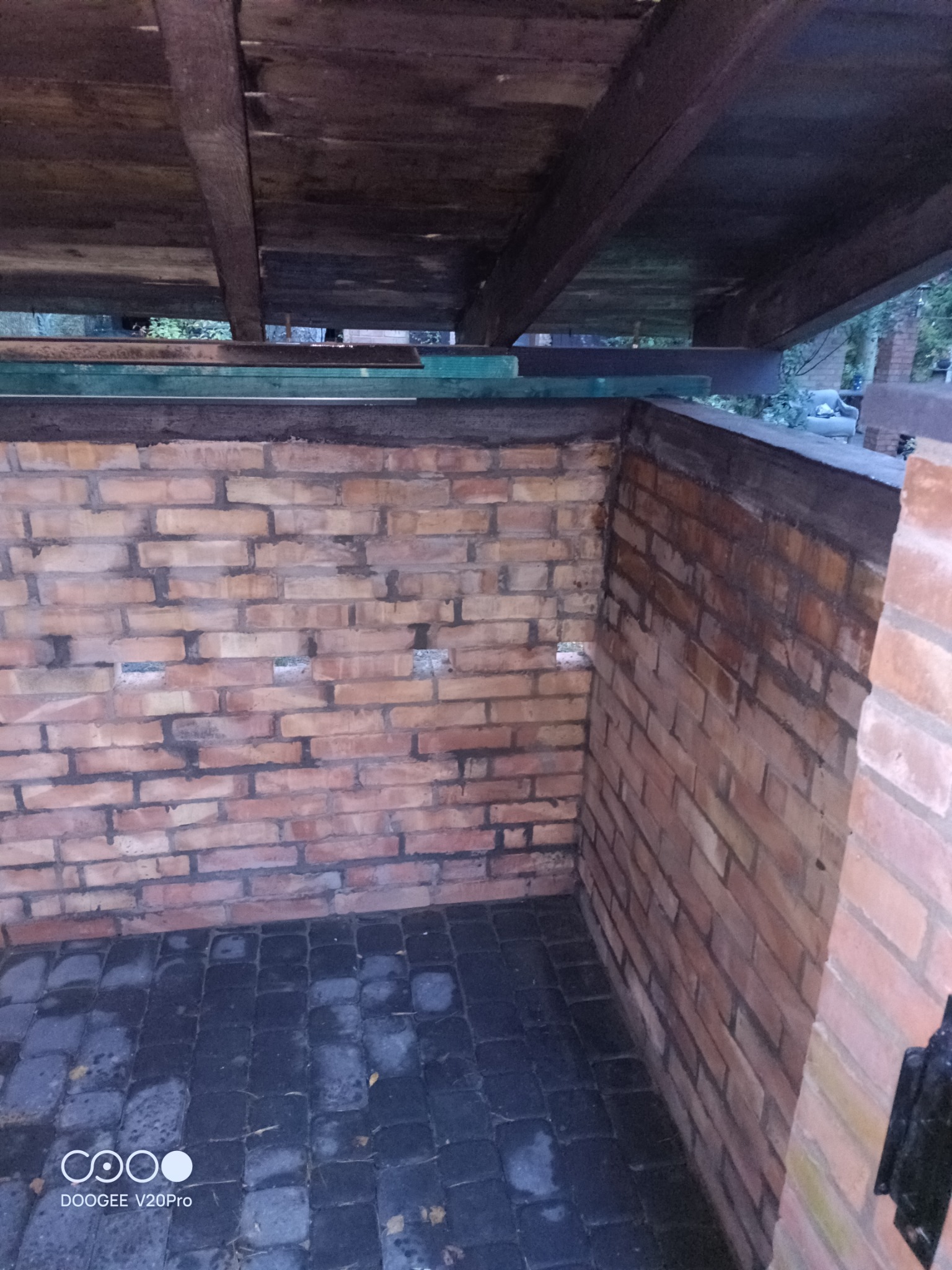 Niedokończona murowana konstrukcja z cegły z drewnianym zadaszeniem, widoczne otwory w murze i ciemna kostka brukowa na podłożu.