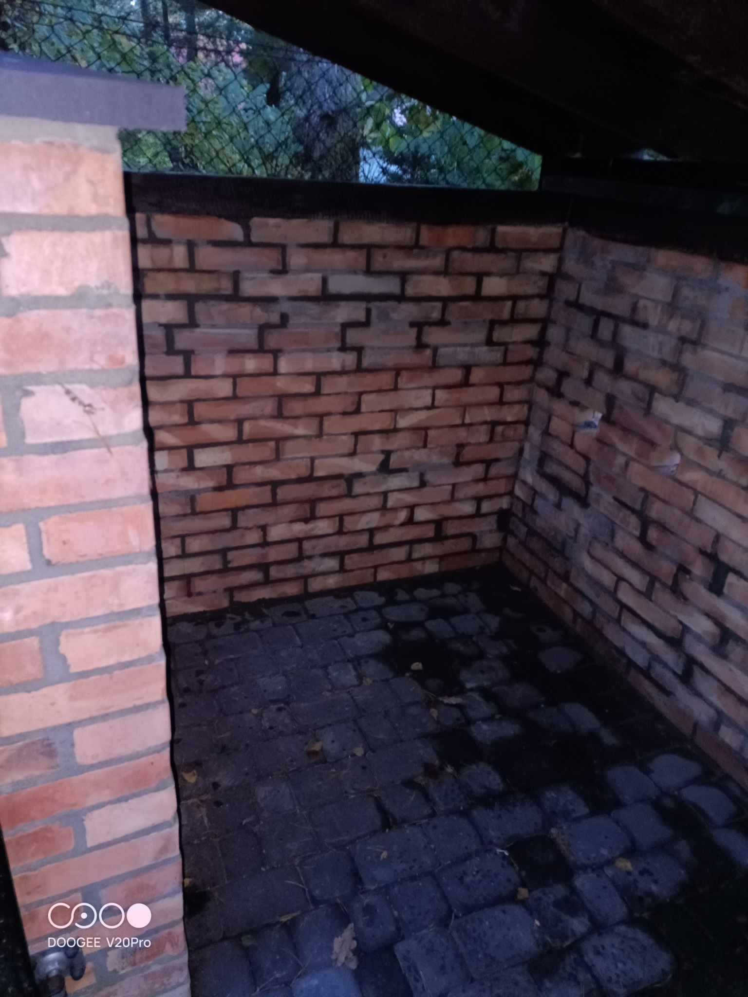 Murowana altana z cegły klinkierowej, wykończona czarną listwą, z podłogą wyłożoną kostką brukową, widok na zewnątrz przez siatkę ogrodzeniową.