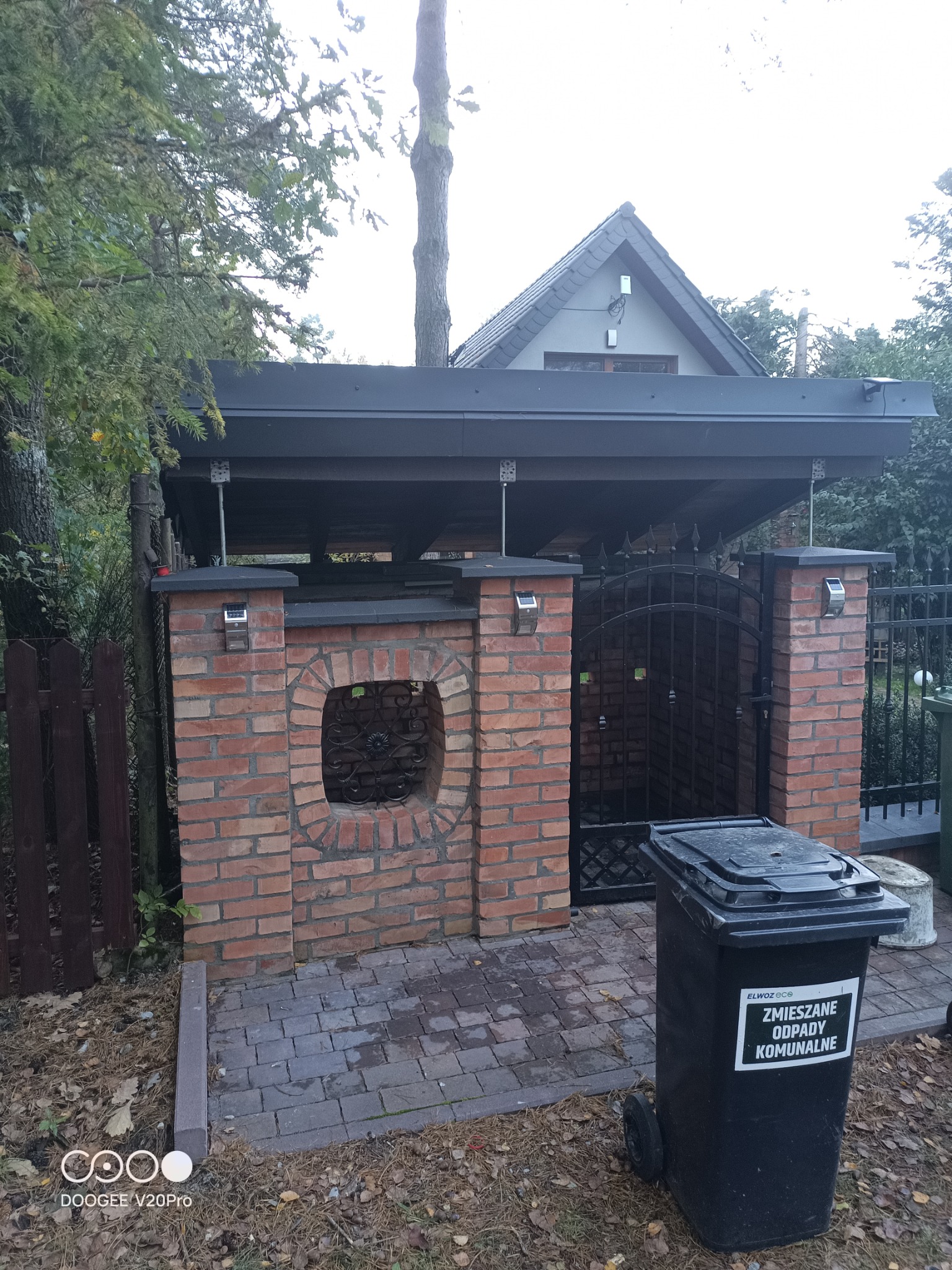Zadaszone miejsce na pojemniki na śmieci z ceglanymi ściankami, ozdobnym okienkiem i metalową bramką, obok drewniany płot i pojemnik na odpady komunalne.