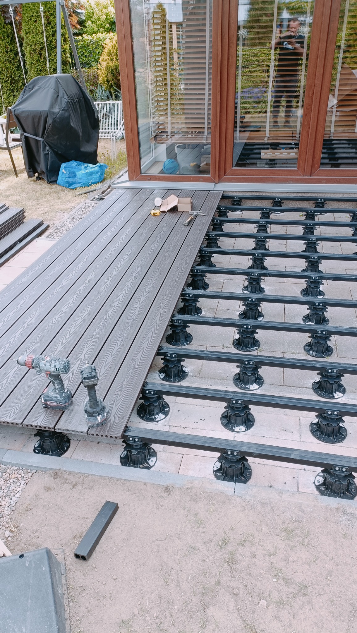 Montaż kompozytowej deski tarasowej na regulowanych wspornikach; widoczne dwa akumulatorowe wkrętarki, miara zwijana i poziomica; w tle okno z żaluzjami.