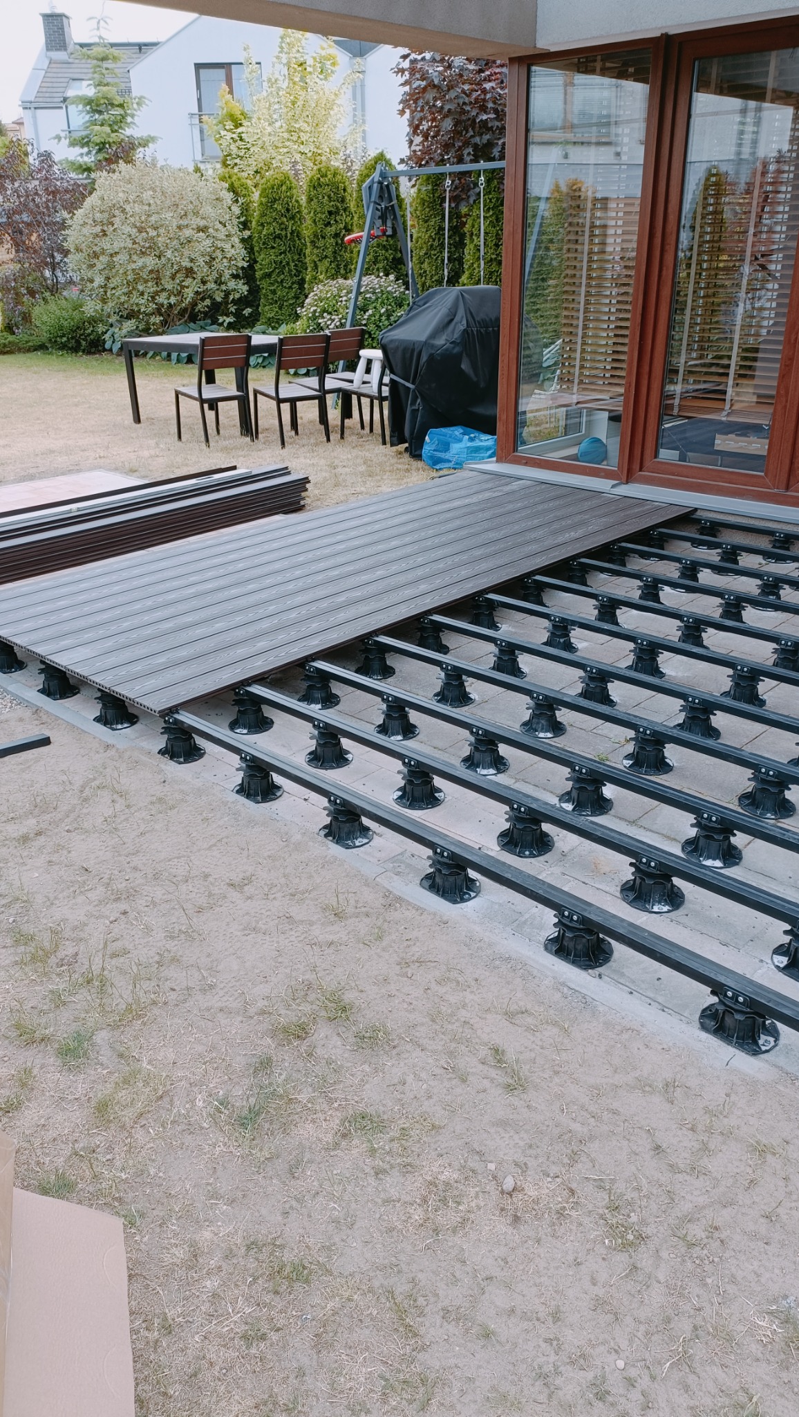 Montaż kompozytowego tarasu: widoczne legary na regulowanych wspornikach i deski tarasowe w kolorze ciemnego drewna, w tle ogród z meblami ogrodowymi i grillem.