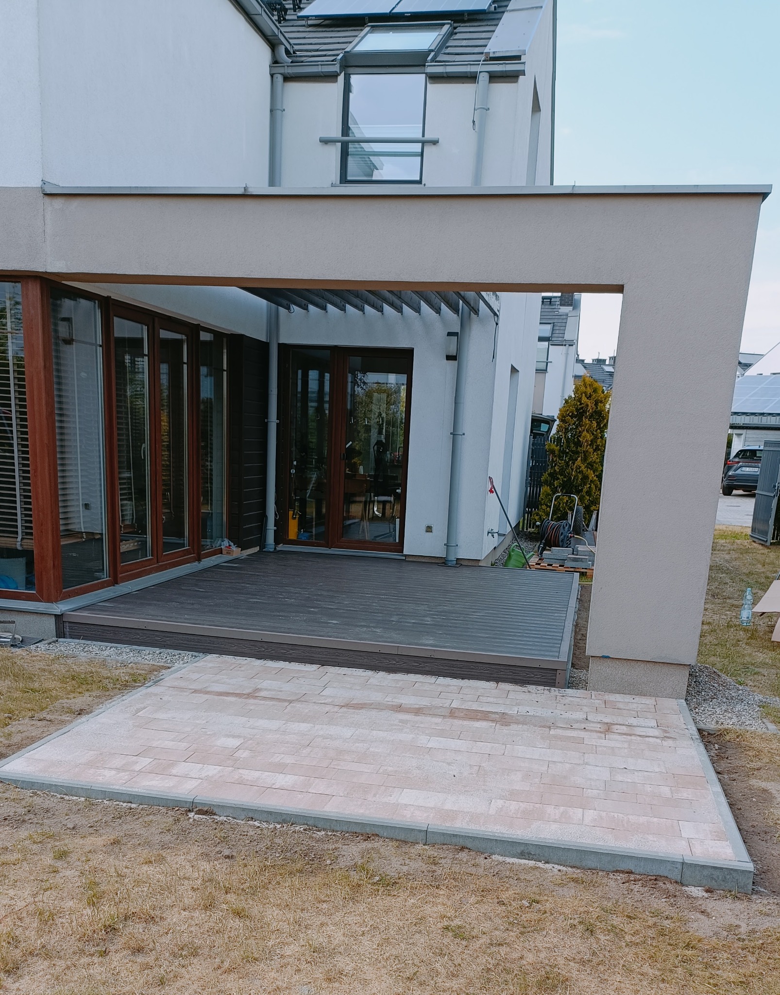 Nowoczesny taras z kompozytowej deski w kolorze grafitowym, połączony z wybrukowanym patio z jasnych płyt, otoczony trawnikiem, na tle białego domu z dużymi oknami.