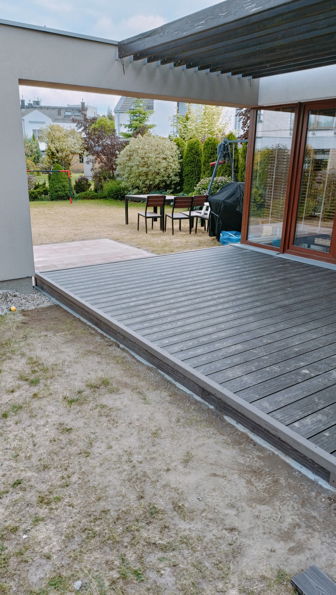 Nowoczesny taras z desek kompozytowych w kolorze szarości, z widokiem na zadbany ogród z meblami ogrodowymi i huśtawką.