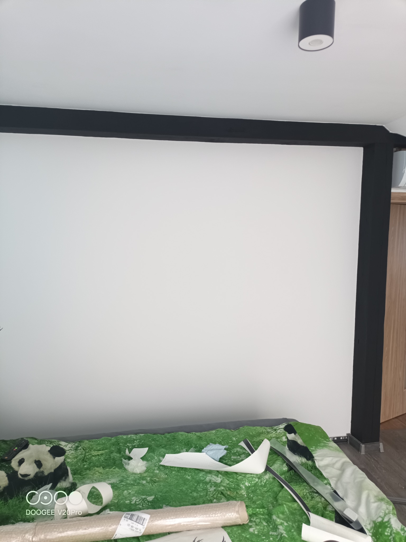 Przygotowania do remontu pokoju: biała ściana z czarnymi elementami konstrukcyjnymi, na łóżku zielona narzuta z motywem trawy, rozłożone elementy dekoracyjne, w tle drewniane drzwi.