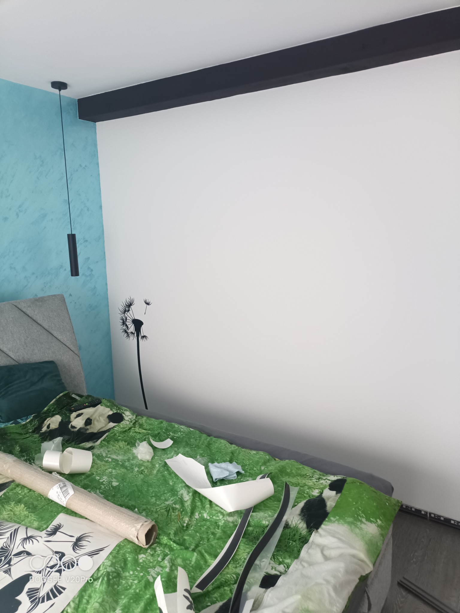 Biała ściana w pokoju z naklejoną czarną naklejką dmuchawca, częściowo naklejone elementy na łóżku, niebieska ściana z efektem dekoracyjnym, czarna lampa wisząca i dekoracyjny element sufitu.