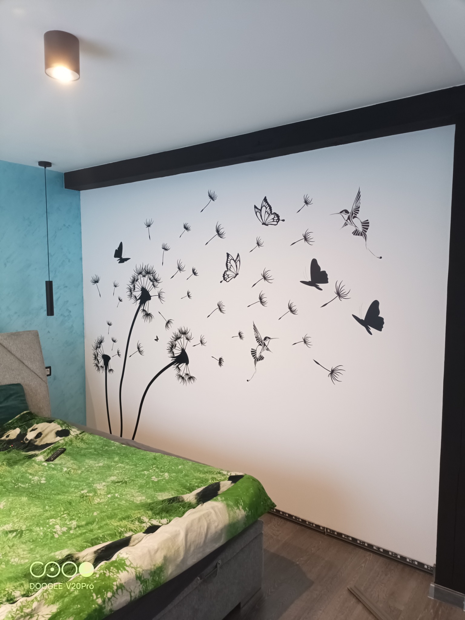 Biała ściana w sypialni z naklejonym wzorem dmuchawców, motyli i kolibrów w kolorze czarnym, widoczne oświetlenie sufitowe i fragment łóżka z zieloną pościelą.