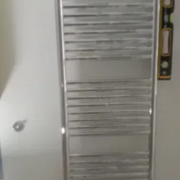 Zamontowany, chromowany grzejnik łazienkowy drabinkowy z poziomnicą przyłożoną z boku, widoczny element instalacji w tle.