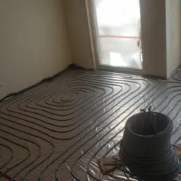 Wężownica ogrzewania podłogowego ułożona na izolacji z siatką, zwój rur w rogu pokoju, okna zabezpieczone folią malarską, widok z góry.