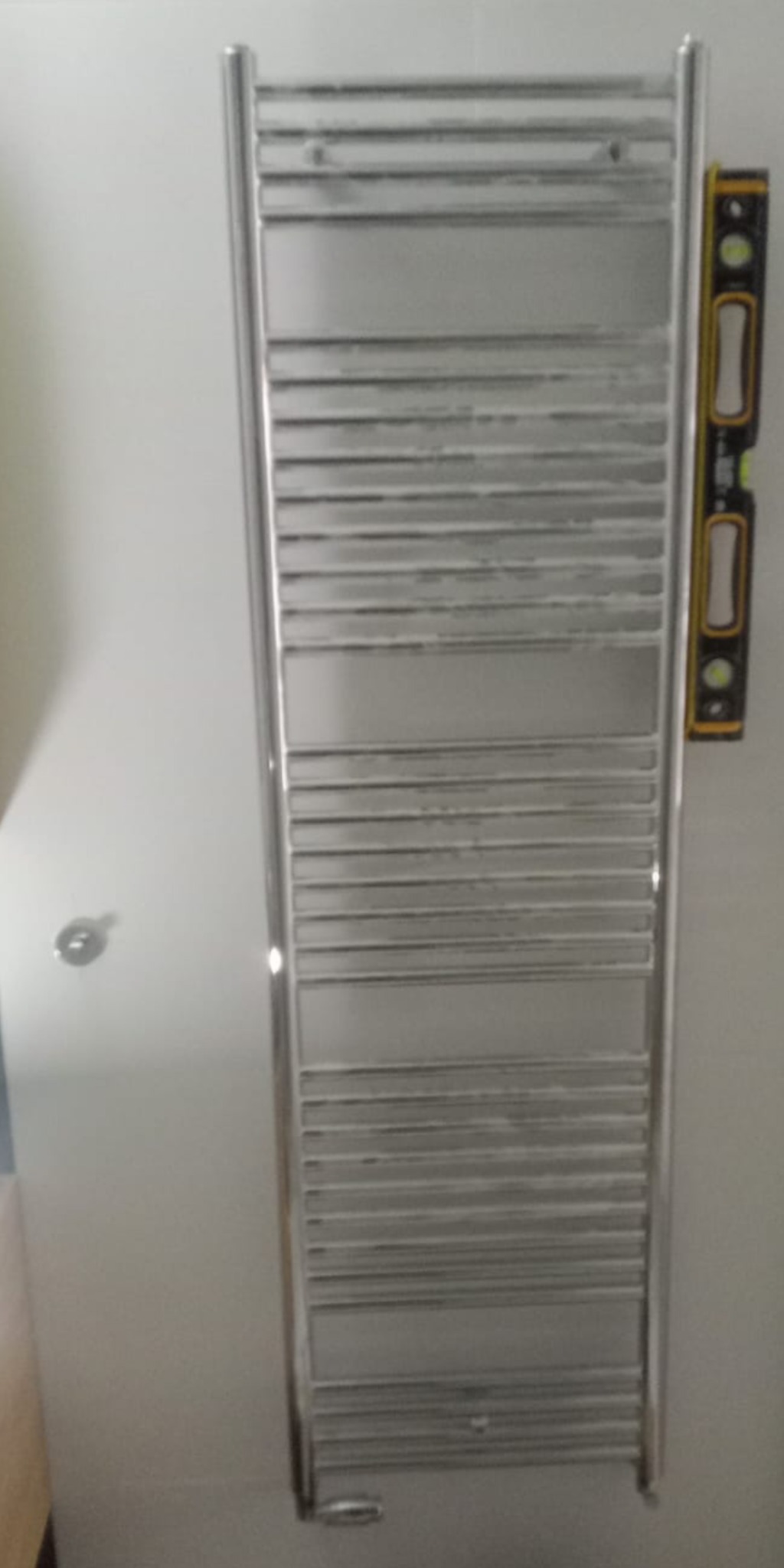 Zamontowany, chromowany grzejnik łazienkowy drabinkowy z poziomnicą przyłożoną z boku, widoczny element instalacji w tle.