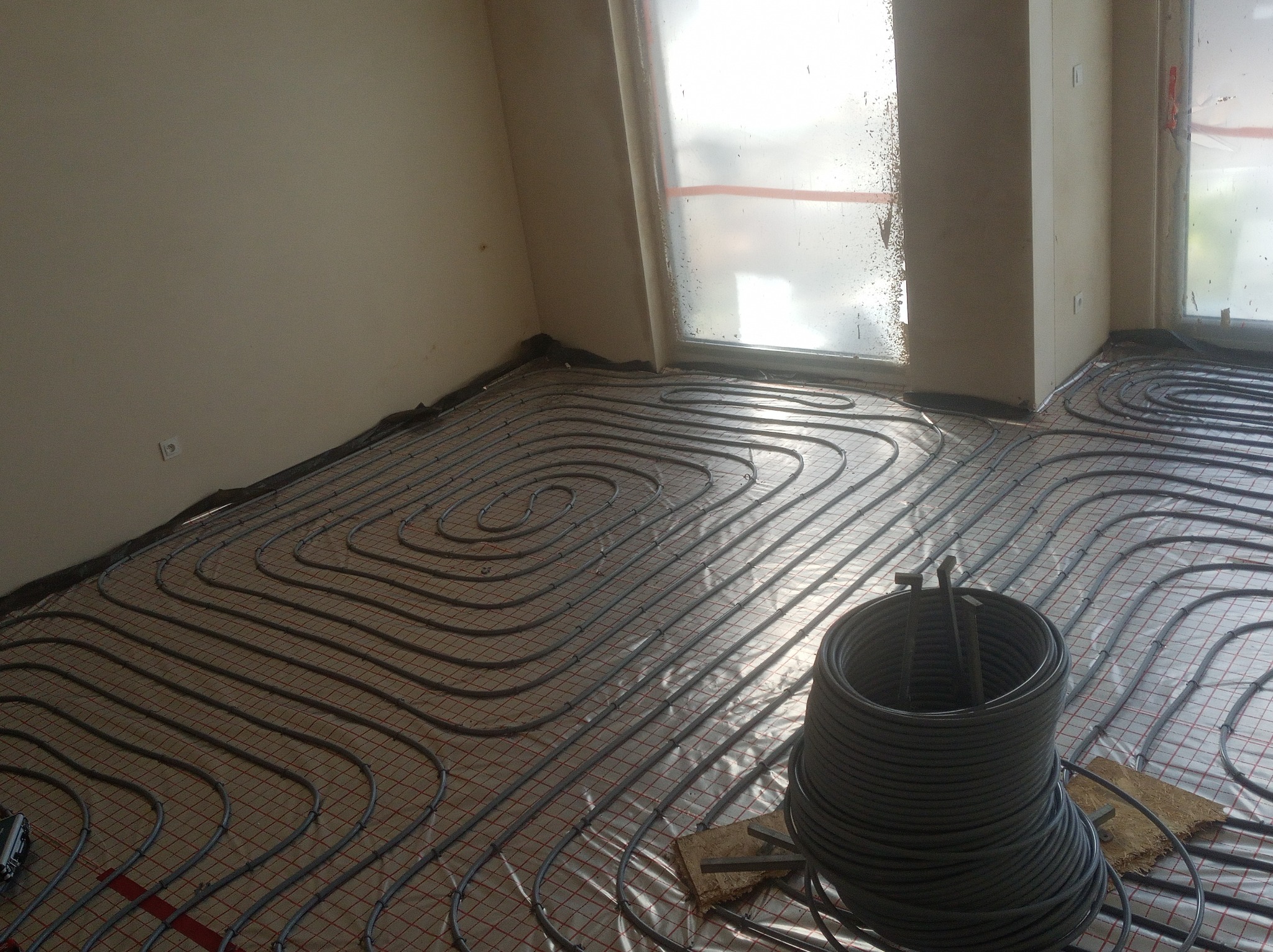 Wężownica ogrzewania podłogowego ułożona na izolacji z siatką, zwój rur w rogu pokoju, okna zabezpieczone folią malarską, widok z góry.