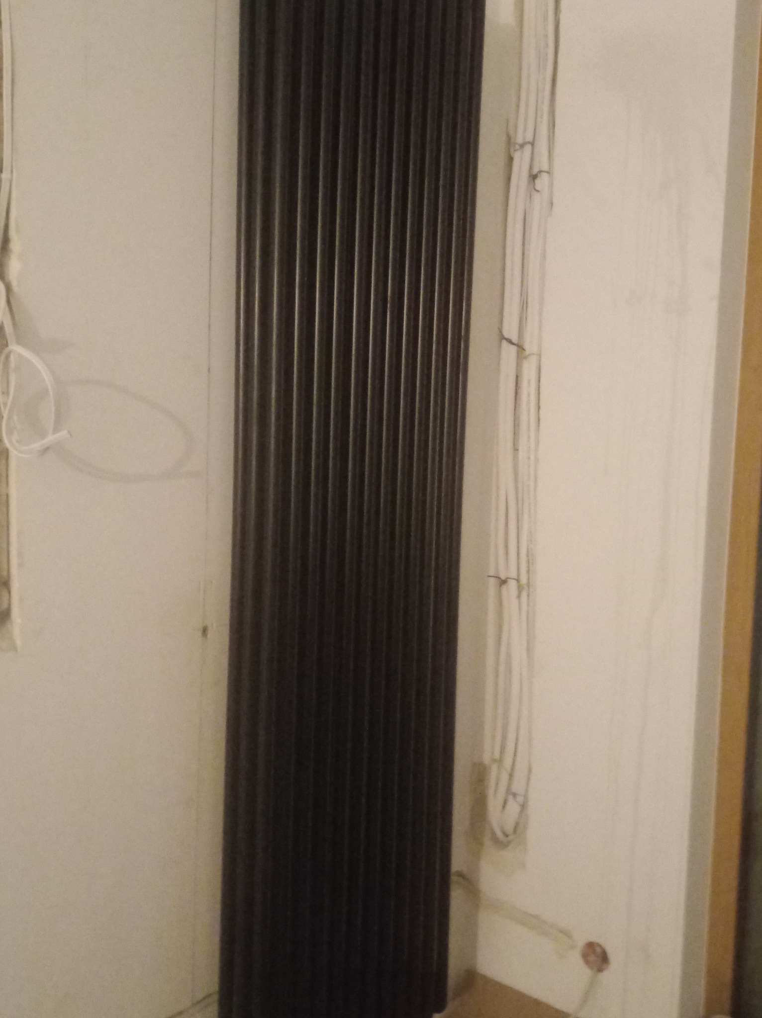 Ciemny, pionowy grzejnik panelowy zamontowany na ścianie. Widoczne wystające kable elektryczne i rurki instalacyjne w ścianie, sugerujące etap prac wykończeniowych.
