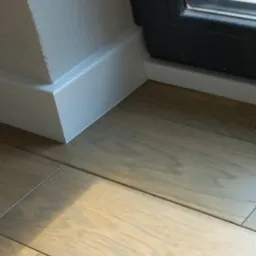 Fragment podłogi z jasnych paneli drewnianych, białe listwy przypodłogowe przy ścianie i czarna rama okienna w tle.