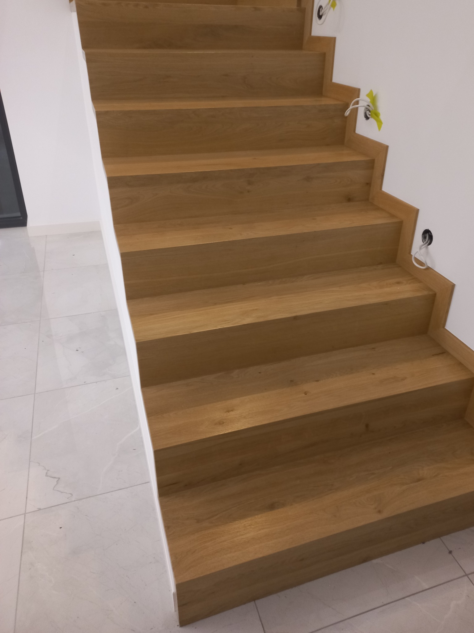 Nowoczesne schody z dębowego drewna, widoczne kable wystające ze ściany w miejscach montażu oświetlenia, białe ściany i podłoga z płytek imitujących marmur.