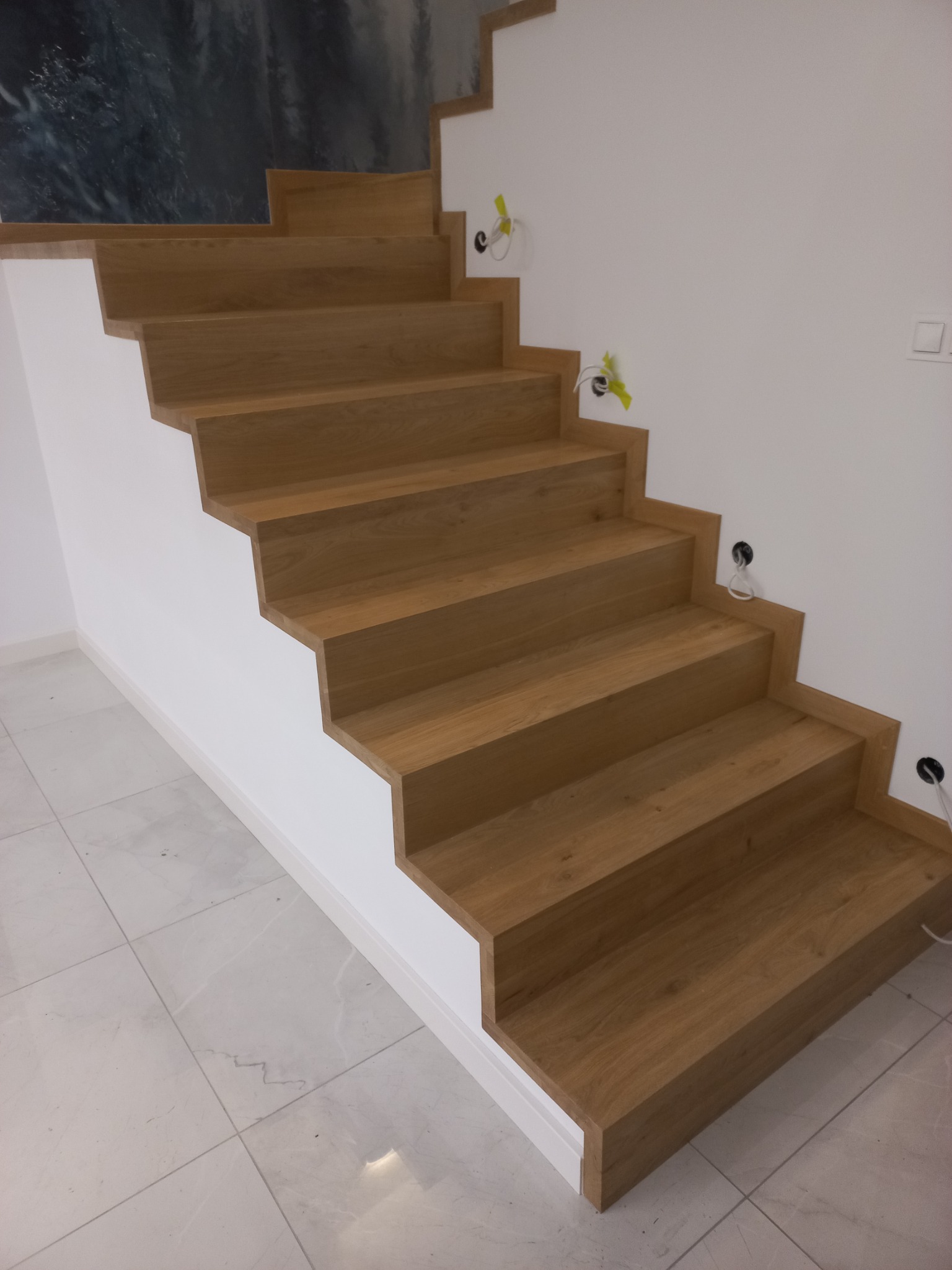 Nowoczesne schody z drewnianymi stopniami o jasnym odcieniu, osadzone na białej konstrukcji, z widocznymi kablami wystającymi ze ściany i marmurową podłogą.