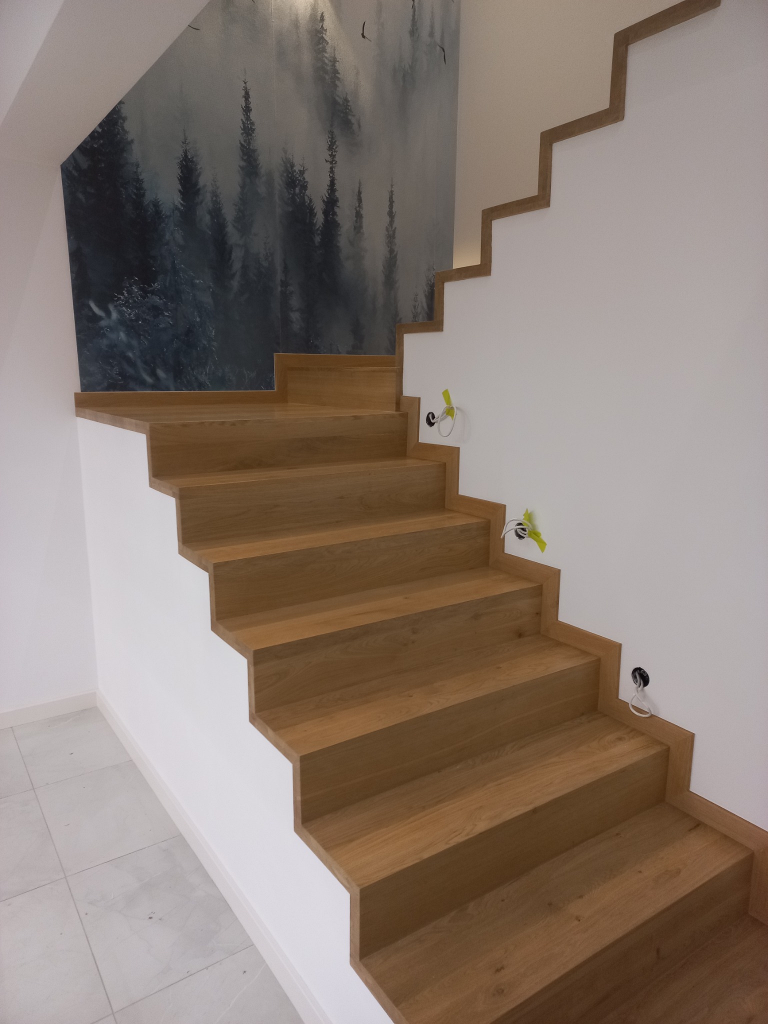 Nowoczesne, dębowe schody z minimalistycznym wykończeniem, zintegrowane z białą ścianą i ozdobną tapetą z motywem leśnym, widoczne przygotowane punkty oświetleniowe.