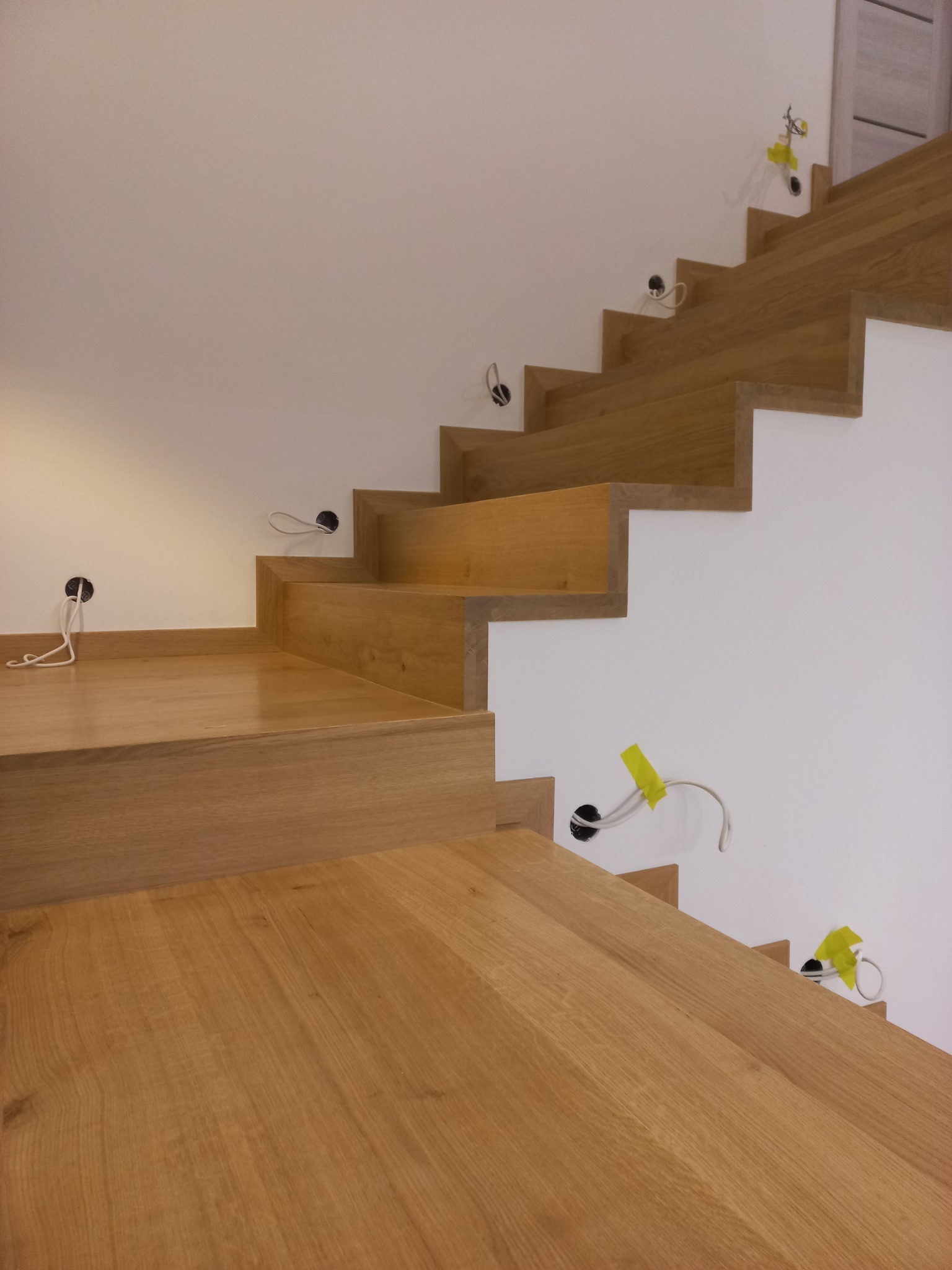 Surowe, dębowe stopnie schodowe w trakcie montażu, z widocznymi przewodami elektrycznymi wystającymi ze ściany, zabezpieczonymi żółtą taśmą.