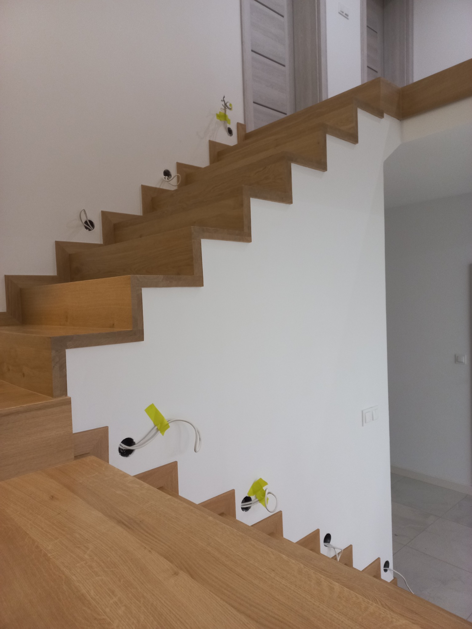 Nowoczesne schody z jasnego drewna, częściowo zamontowane, z widocznymi przewodami elektrycznymi wystającymi ze ściany, zabezpieczonymi żółtą taśmą.