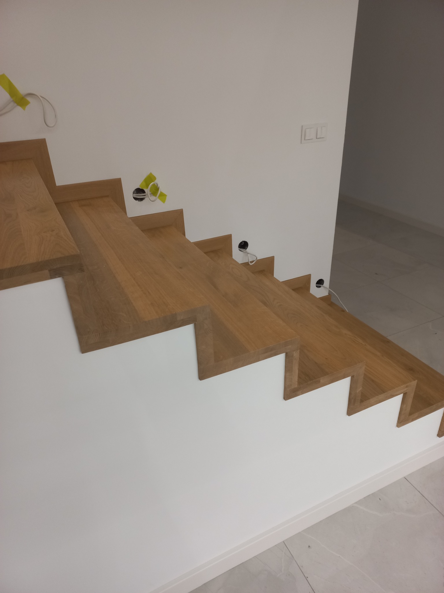 Stopnie w trakcie montażu, widoczne otwory w ścianie na kable i taśma malarska, drewno w ciepłym odcieniu, biała ściana.