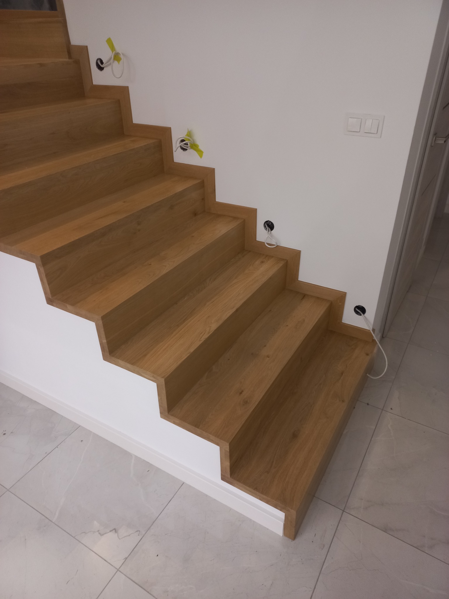 Nowoczesne, dębowe stopnie schodów w trakcie montażu, widoczne wystające kable elektryczne i puszki instalacyjne na białej ścianie, marmurowa podłoga.