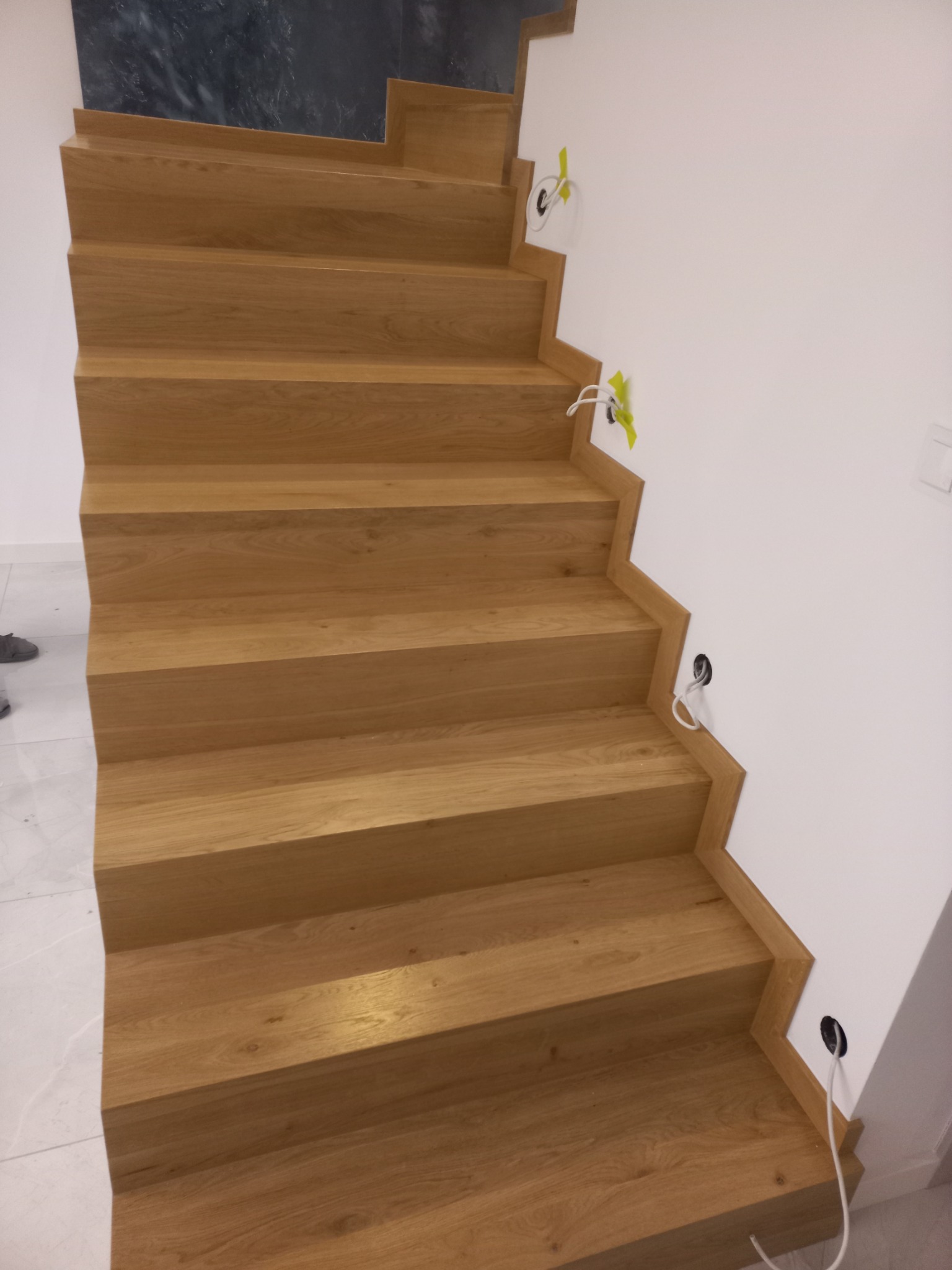 Klatka schodowa z jasnego drewna w trakcie montażu, widoczne wystające kable elektryczne i taśma maskująca.