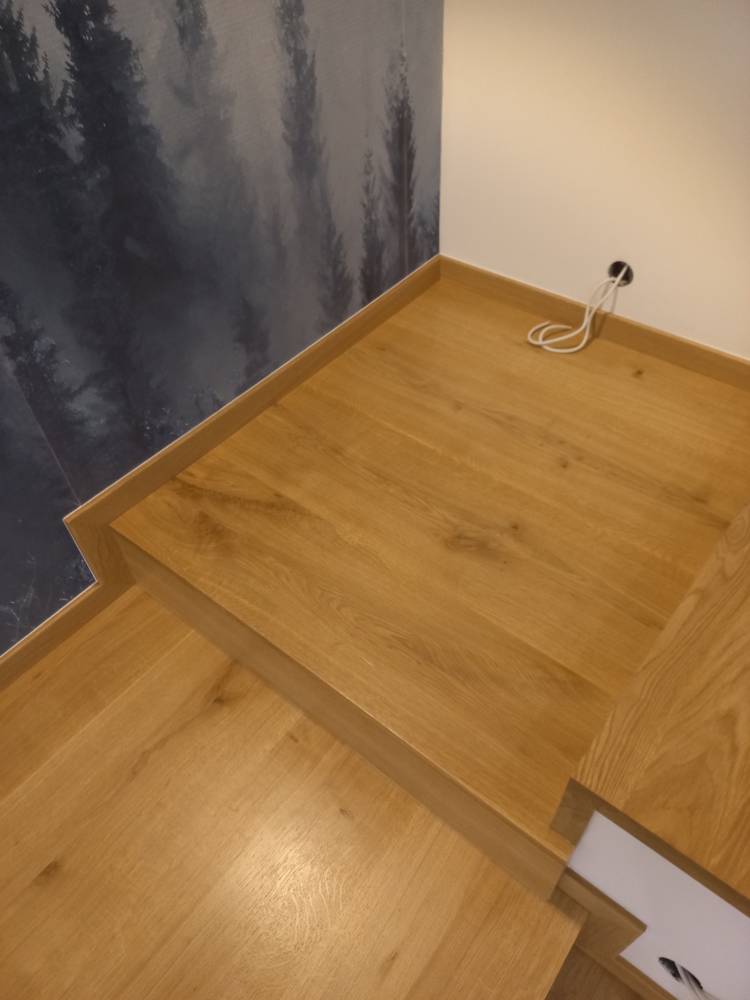 Fragment drewnianych schodów z dębowymi stopniami, widoczne okablowanie przy ścianie i tapeta z motywem leśnym.