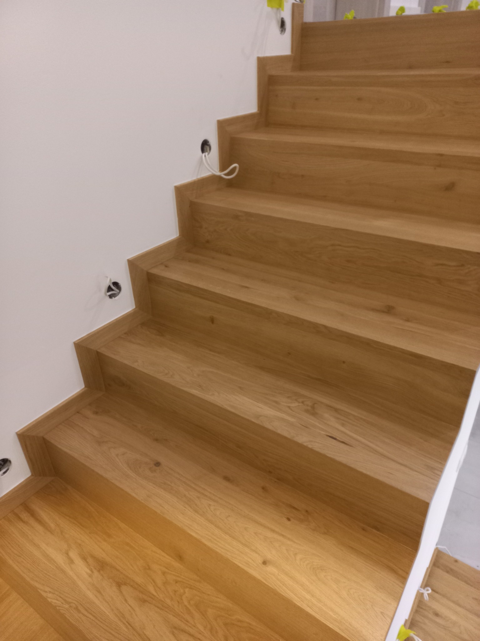 Nowoczesne, dębowe schody z minimalistycznym wykończeniem, widoczne otwory na oświetlenie w ścianie, zabezpieczone folią ochronną na górnych stopniach.