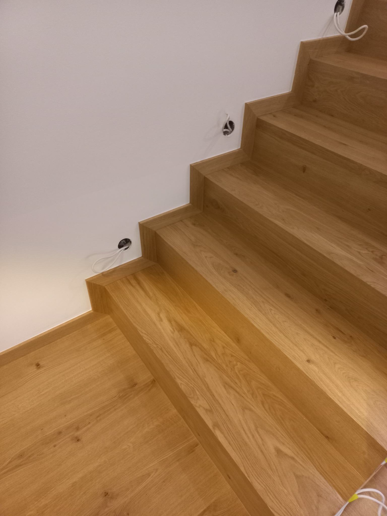 Widok z góry na dębowe stopnie schodów z podświetleniem w ścianie, widoczne otwory instalacyjne przed montażem lamp.