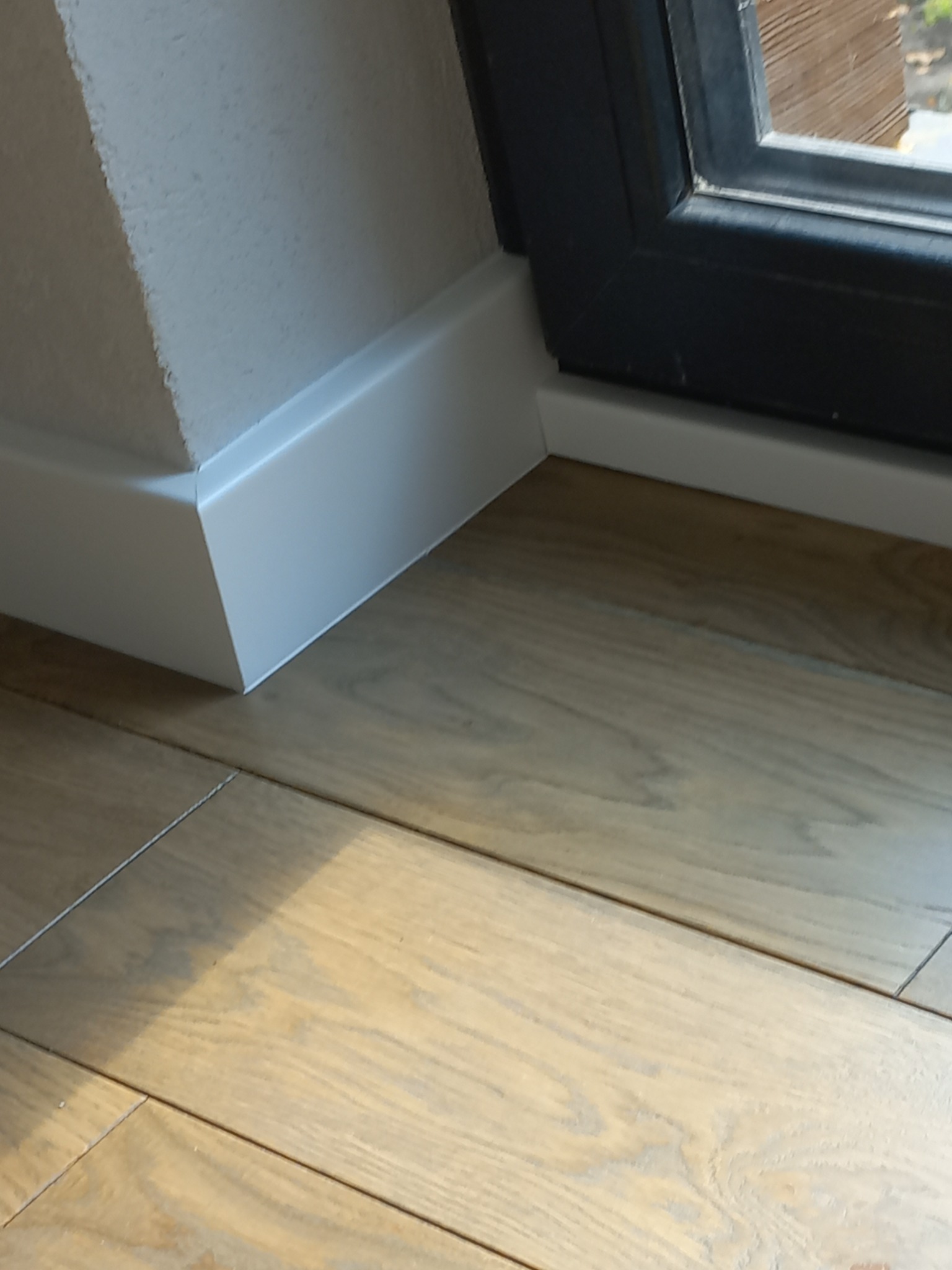 Fragment podłogi z jasnych paneli drewnianych, białe listwy przypodłogowe przy ścianie i czarna rama okienna w tle.