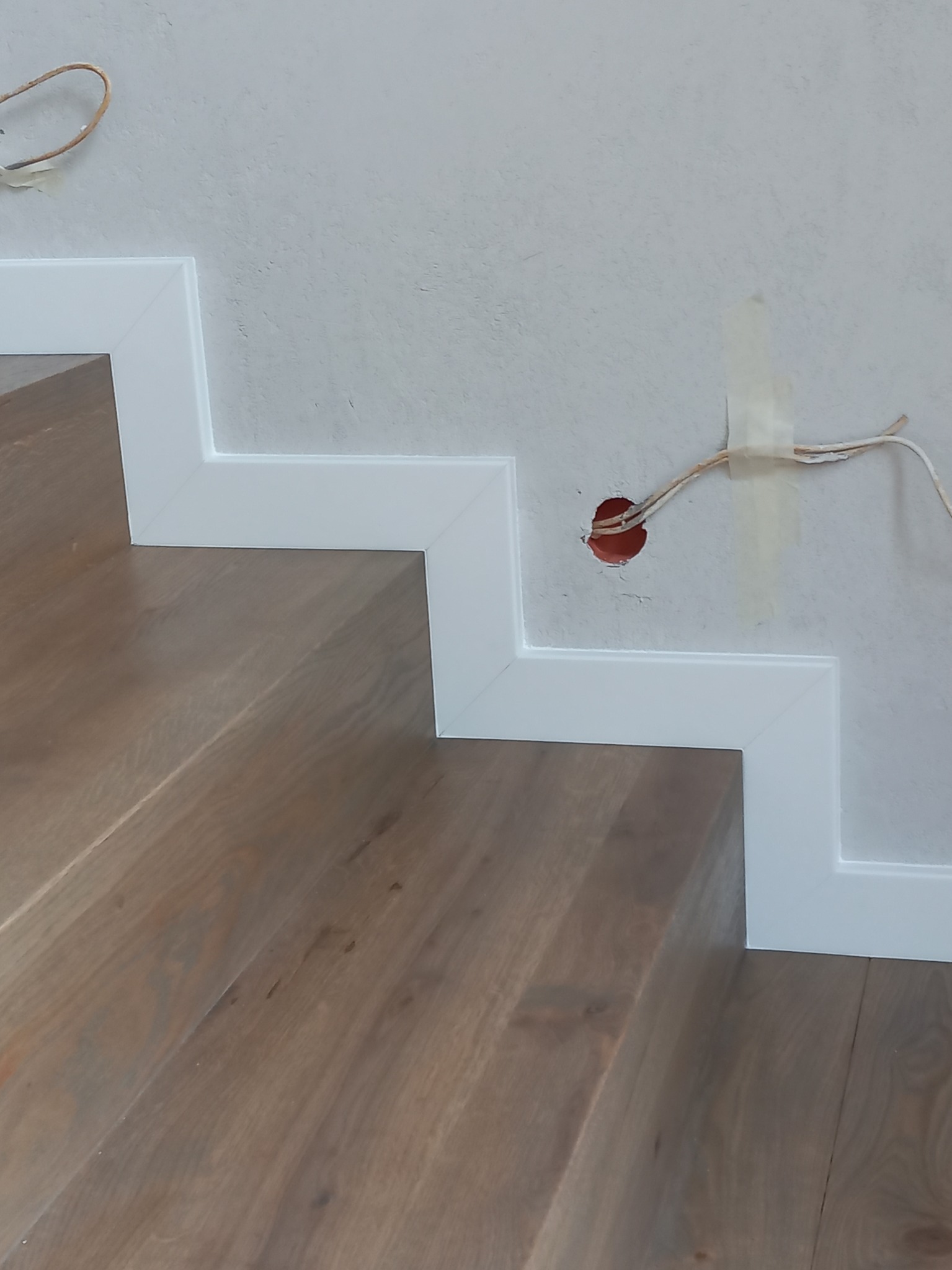 Fragment drewnianych schodów z białym wykończeniem przy ścianie, widoczny otwór w ścianie z wystającymi kablami.