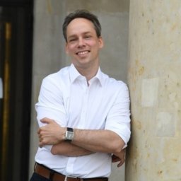 Przemysław Wojewoda - Uśmiechnięty mężczyzna w białej koszuli oparty o marmurową kolumnę, z zegarkiem na ręku, w tle przeszklone drzwi.