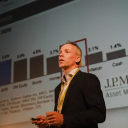 Prelegent w garniturze prezentuje wykresy inwestycyjne na tle ekranu podczas konferencji finansowej. Widoczny wskaźnik inflacji i logo J.P. Morgan Asset Management.