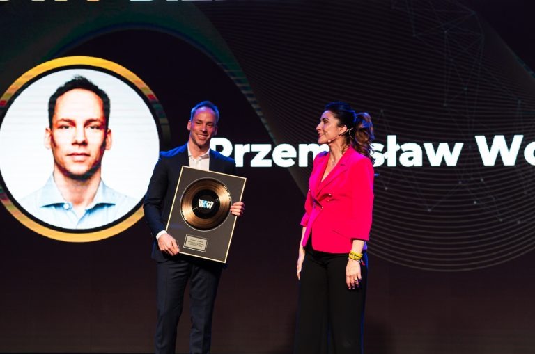 Mężczyzna w garniturze otrzymuje nagrodę WOW z logo Oferteo na scenie, obok stoi kobieta w różowej marynarce.