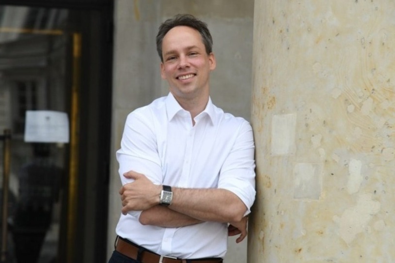 Uśmiechnięty mężczyzna w białej koszuli oparty o marmurową kolumnę, z zegarkiem na ręku, w tle przeszklone drzwi.