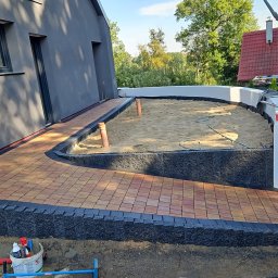 ProBudTech WOJCIECH GEMZA - Ułożona kostka brukowa w odcieniach czerwieni i brązu, obramowana czarnym obrzeżem, prowadząca do obszaru wypełnionego piaskiem z wystającymi rurami PCV, widoczne narzędzia i materiały budowlane...