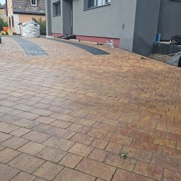 ProBudTech WOJCIECH GEMZA - Nowo ułożona, brązowo-pomarańczowa kostka brukowa na podjeździe do nowoczesnego domu z szarą elewacją, z widocznymi workami z materiałem budowlanym i częściowo ukrytą maszyną do zagęszczania gruntu.