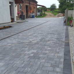 ProBudTech WOJCIECH GEMZA - Ułożona szara kostka brukowa na podjeździe z liniowym odpływem, obok rabata kwiatowa i pas z białym grysem przy ścianie budynku.