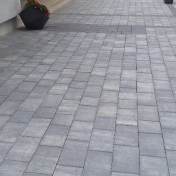 ProBudTech WOJCIECH GEMZA - Ułożona szara kostka brukowa na podjeździe przed domem z widocznymi donicami z kwiatami i częścią ogrodu w tle.