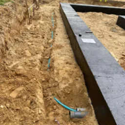 Wykop pod fundamenty z izolacją i niebieskimi rurami instalacji wodnej w ziemi. Widoczne narzędzia pomiarowe i dokumentacja.