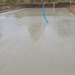 ProBudTech WOJCIECH GEMZA - Wylana, gładka płyta fundamentowa z wystającą niebieską rurą instalacyjną. W tle niwelacja terenu i drzewa.