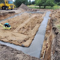 ProBudTech WOJCIECH GEMZA - Wykop pod fundamenty z wylaną ławą fundamentową, ubijarką i niwelatorem laserowym na placu budowy. W tle żółty dźwig samochodowy i hałdy ziemi.